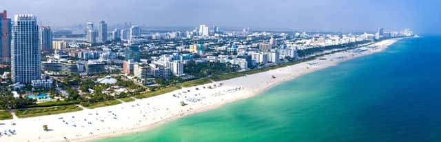 Una vista aerea di un moderno paesaggio urbano con grattacieli lungo un'ampia spiaggia di sabbia bianca, accanto all'acqua turchese dell'oceano.