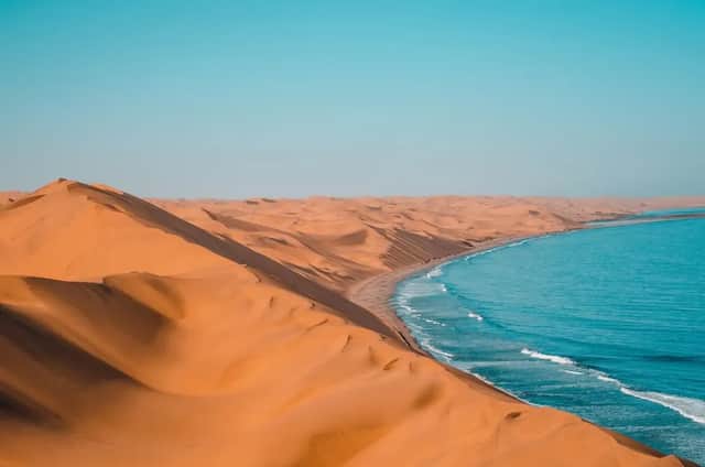 Weite orangefarbene Sanddünen treffen auf den blauen Ozean und bilden eine Küstenlinie unter klarem Himmel.