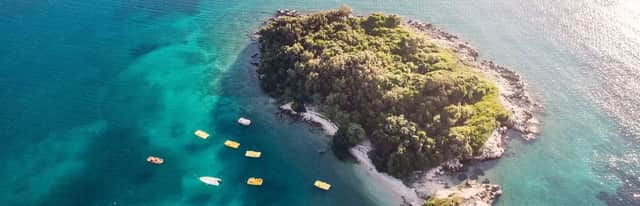 Luftaufnahme einer kleinen, bewaldeten Insel in türkisfarbenem Wasser, mit mehreren gelben Tretbooten in der Nähe.