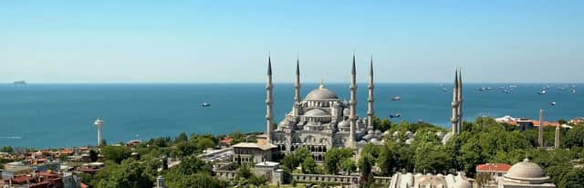 A large mosque with multiple domes and six minarets overlooking a sea filled with ships under a clear blue sky.