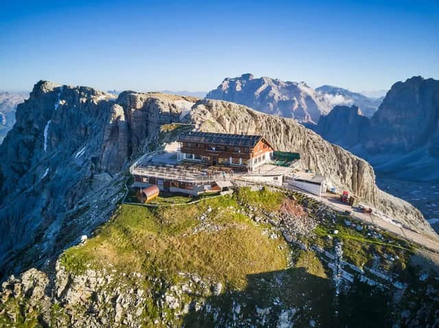 Una vista aerea di un rifugio alpino in legno con terrazza, arroccato su una cima rocciosa sotto un cielo azzurro.