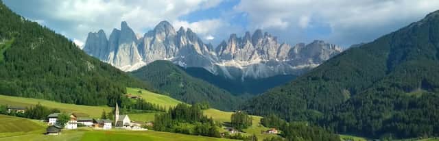 Un piccolo villaggio con una chiesa sorge in una verde valle ai piedi di colline boscose e di una catena montuosa rocciosa e frastagliata.