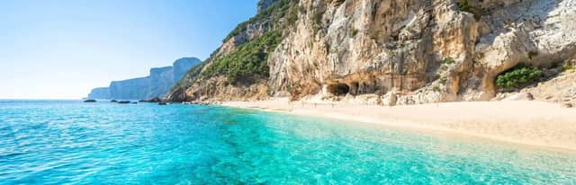 Acque turchesi lambiscono una spiaggia sabbiosa e appartata ai piedi di un'imponente scogliera frastagliata sotto un cielo limpido.