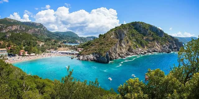 Una vista elevada de una playa concurrida con agua turquesa en una cala, rodeada de acantilados cubiertos de árboles y montañas.