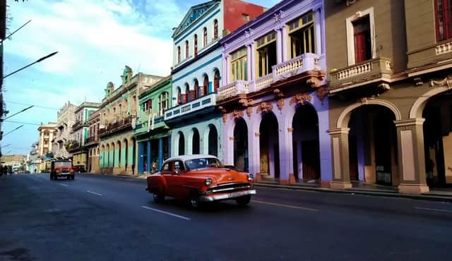 Un coche vintage naranja circula por una calle junto a coloridos edificios coloniales con puertas arqueadas bajo un cielo azul.