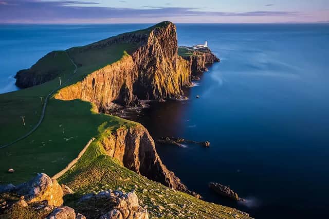 Un faro blanco se asienta en una península rocosa y cubierta de hierba que se adentra en el tranquilo mar azul, iluminado por la cálida luz del atardecer.