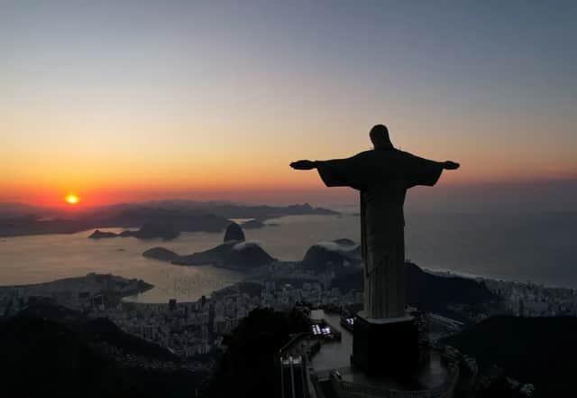 The silhouette of a large statue with outstretched arms overlooks a coastal city, a bay, and mountains during a colorful sunrise.