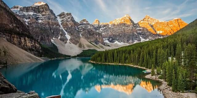 Montagne innevate illuminate dal sole si riflettono in un lago turchese calmo circondato da una fitta foresta di pini.