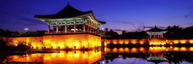 Una tradizionale pagoda asiatica e un muro di pietra illuminati al tramonto, con le loro luci calde che si riflettono nell'acqua immobile.