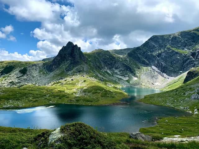 Un lac d'un bleu profond est niché dans une vallée de montagne verdoyante, avec des sommets rocheux sous un ciel aux nuages cotonneux.