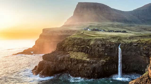 Una cascata si getta da una verde scogliera erbosa nell'oceano, con un villaggio sullo sfondo al tramonto.