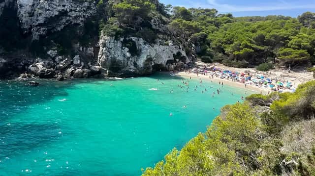 Veduta dall'alto di una spiaggia affollata in una cala rocciosa, con persone che nuotano nelle acque turchesi e si rilassano sulla sabbia.