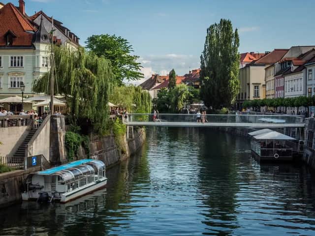 Un moderno puente de cristal cruza un río urbano con barcos turísticos. Las orillas están bordeadas de edificios históricos, árboles y cafeterías al aire libre.