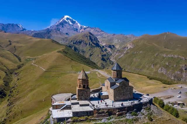 Una vista aerea di un monastero di pietra su una collina, con una vetta montana innevata che si staglia in lontananza su colline verdeggianti.