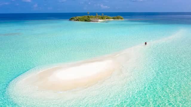 Una vista aerea di due persone che camminano su un banco di sabbia bianca che si estende attraverso acque cristalline color turchese verso una piccola isola tropicale.