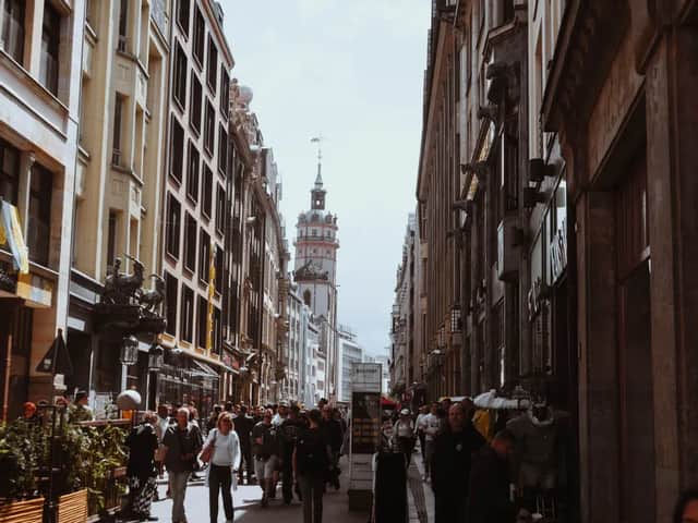 Eine Menschenmenge geht eine enge Stadtstraße entlang, die von hohen, historischen Gebäuden und einem fernen Glockenturm gesäumt ist.