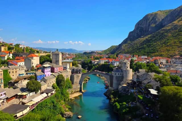 Eine alte Steinbrücke wölbt sich über einen türkisfarbenen Fluss, der durch eine historische Stadt mit Hangbebauung unter einem klaren blauen Himmel fließt.