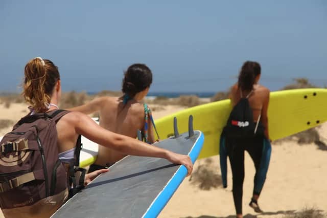 Un viaggio di gruppo WeRoad con tre donne viste di spalle, che trasportano tavole da surf mentre camminano su un sentiero sabbioso verso il mare.