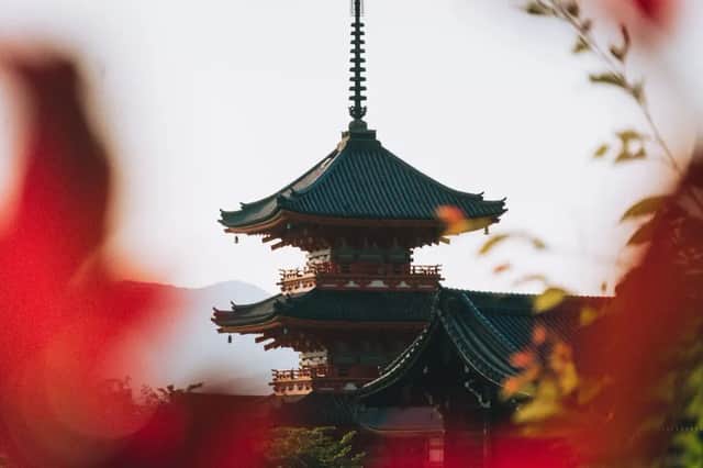 Eine mehrstöckige japanische Pagode mit einem dunklen Ziegeldach ist durch verschwommene rote Blätter vor einer dunstigen Bergkulisse zu sehen.
