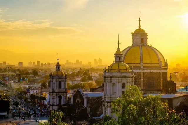 Eine historische Kathedrale mit goldenen Kuppeln überblickt eine Stadt mit moderner Skyline während eines goldenen Sonnenuntergangs.