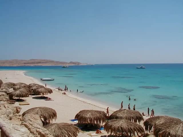 Blick von oben auf einen tropischen Strand mit Strohschirmen, Badenden im türkisfarbenen Wasser und Booten in der Ferne.