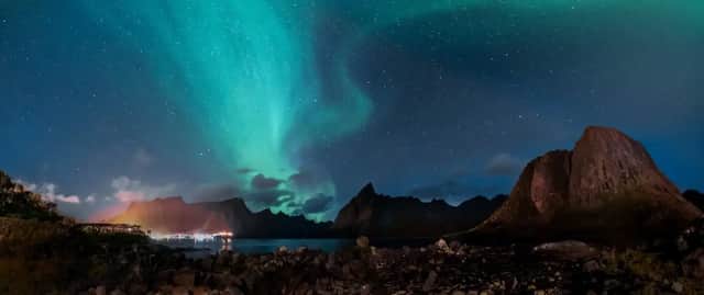L'aurora boreale verde risplende in un cielo notturno stellato su un fiordo, con montagne stagliate e una costa rocciosa sottostante.