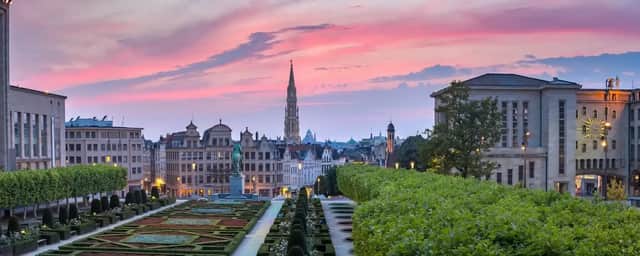Una vista panoramica su un giardino formale verso lo skyline di una storica città europea con un'alta guglia sotto un vivace tramonto rosa.