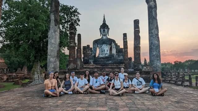 Un gruppo WeRoad si siede a gambe incrociate per una foto davanti a una grande statua di Buddha tra antiche rovine di templi al tramonto.