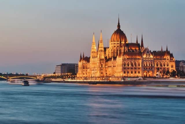 Un grand édifice orné d'un dôme central est illuminé de lumières dorées le long d'une rivière au crépuscule.
