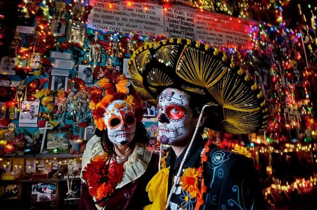 Dos personas con disfraces del Día de Muertos y maquillaje de calavera de azúcar posan frente a una pared decorada con luces y fotos.