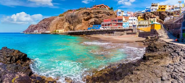 A panoramic view of a coastal village with colorful houses built into a cliff above a turquoise sea and sandy beach.