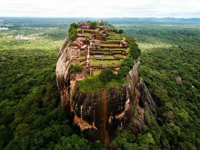 Aerial view of a large rock fortress with ancient ruins on its summit, surrounded by a vast, dense green jungle.