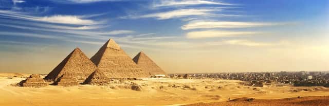 A panoramic view of three large pyramids in a sandy desert with a sprawling city in the distance under a blue, cloudy sky.