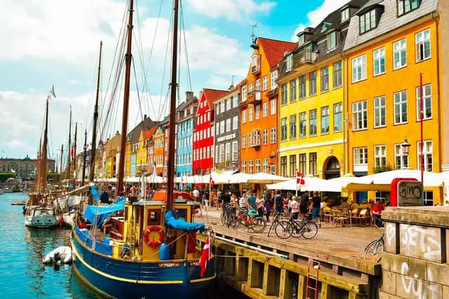 Edificios coloridos bordean un canal donde hay veleros atracados, con gente caminando y sentada en cafeterías a lo largo del muelle frente al mar.