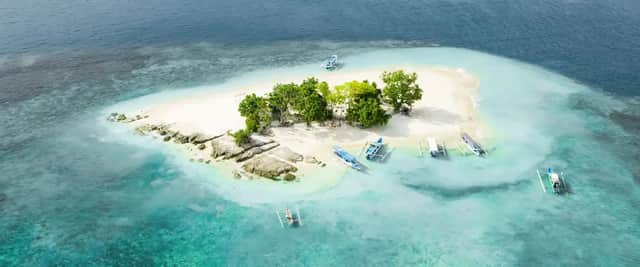 Vista aérea de una pequeña isla tropical con playa de arena blanca, árboles verdes y varios barcos en agua turquesa.