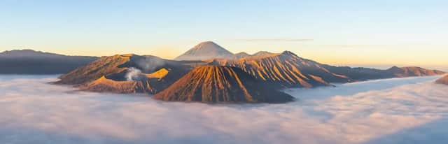 Un volcan fumant et d'autres sommets montagneux s'élèvent au-dessus d'une dense couche de nuages, illuminés par la lumière dorée du lever du soleil.