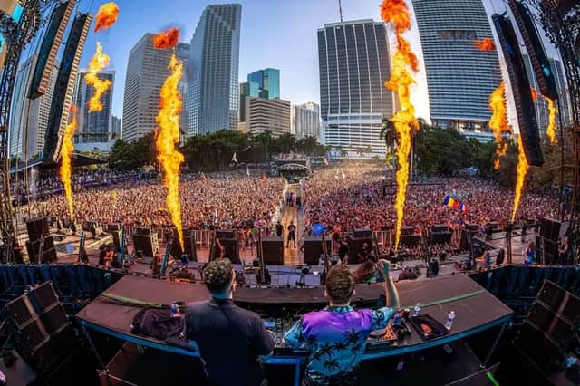 Una vista da dietro due DJ sul palco che guardano una folla immensa a un festival, con fiamme pirotecniche e grattacieli.