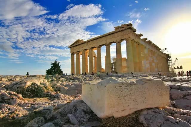 Ein antiker griechischer Tempel mit großen Säulen steht auf einem felsigen Hügel unter einem blauen Himmel mit weißen Wolken bei Sonnenuntergang.