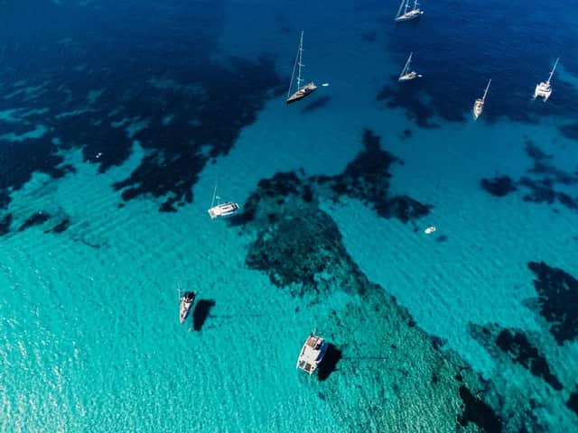 Eine Luftaufnahme von mehreren Segelbooten und Katamaranen, die in transparentem türkisfarbenem Wasser über einem sandigen und felsigen Meeresboden schwimmen.