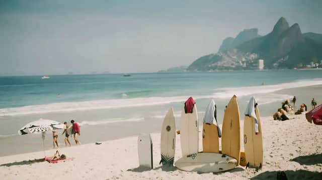Plusieurs planches de surf sont dressées dans le sable sur une plage ensoleillée, avec des gens qui se détendent au bord de l'océan et des montagnes en arrière-plan.