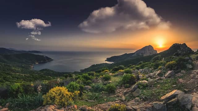 Una costa rocciosa e verde scende verso il mare sotto un cielo nuvoloso al tramonto.