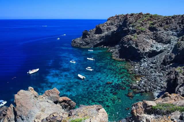Una vista aerea di diverse barche che galleggiano su acqua limpida e turchese all'interno di una caletta rocciosa, sotto un cielo azzurro brillante.