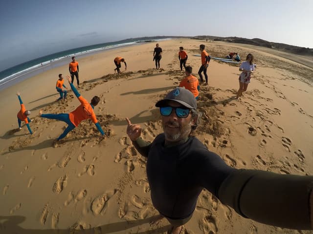Instructor de surf se hace un selfie en la playa mientras un grupo practica en la arena.
