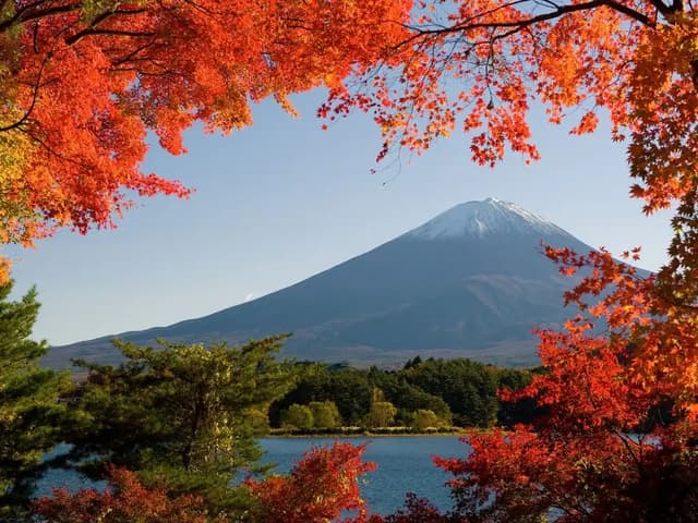 Una montagna innevata incorniciata da vivaci foglie rosse autunnali, che domina un lago circondato da alberi verdi.