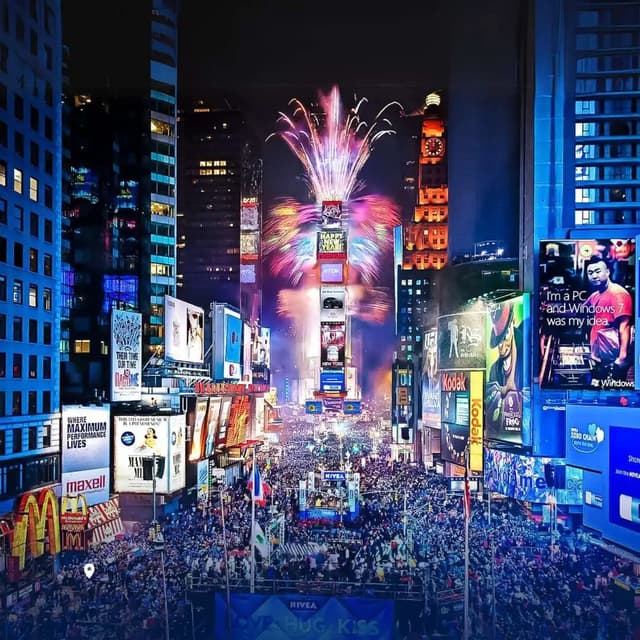 Una folla immensa riempie una piazza di notte, osservando fuochi d'artificio colorati esplodere tra grattacieli illuminati da cartelloni pubblicitari.