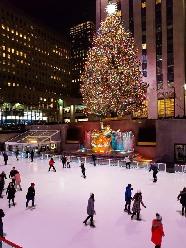 Gente patinando sobre hielo en una pista al aire libre por la noche, con un gran árbol de Navidad iluminado y una estatua dorada de fondo.