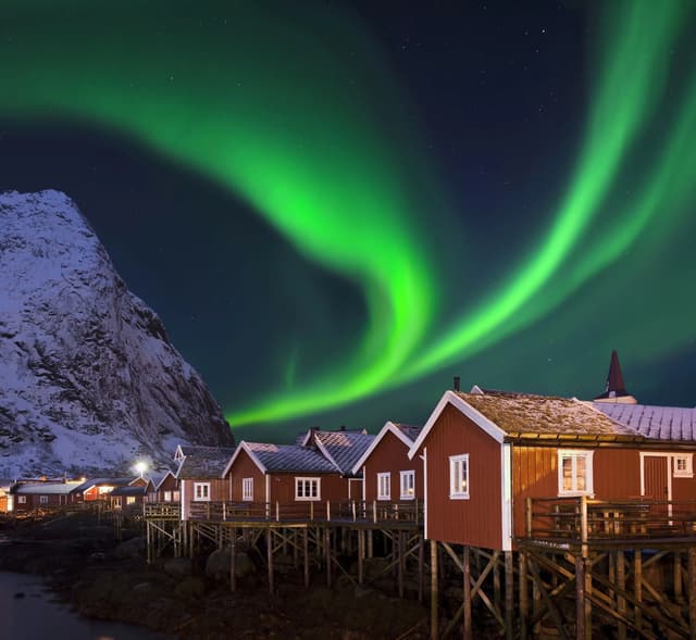 Die grünen Nordlichter leuchten am Sternenhimmel über einem Dorf mit roten Pfahlbauten neben einem verschneiten Berg.