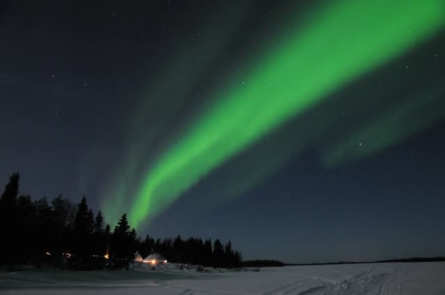 Une aurore boréale verte zèbre un ciel nocturne étoilé au-dessus d'un paysage enneigé avec une ligne d'arbres et de petites cabanes.