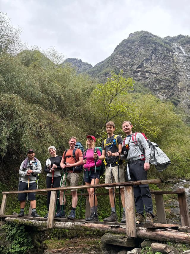 Six randonneurs du groupe WeRoad, équipés de sacs à dos et de bâtons de marche, posant sur un pont en bois au cœur d'une forêt montagnarde.