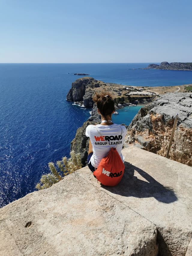 Un chef de groupe WeRoad est assis sur une falaise rocheuse, contemplant la mer d'un bleu profond et un littoral lointain.